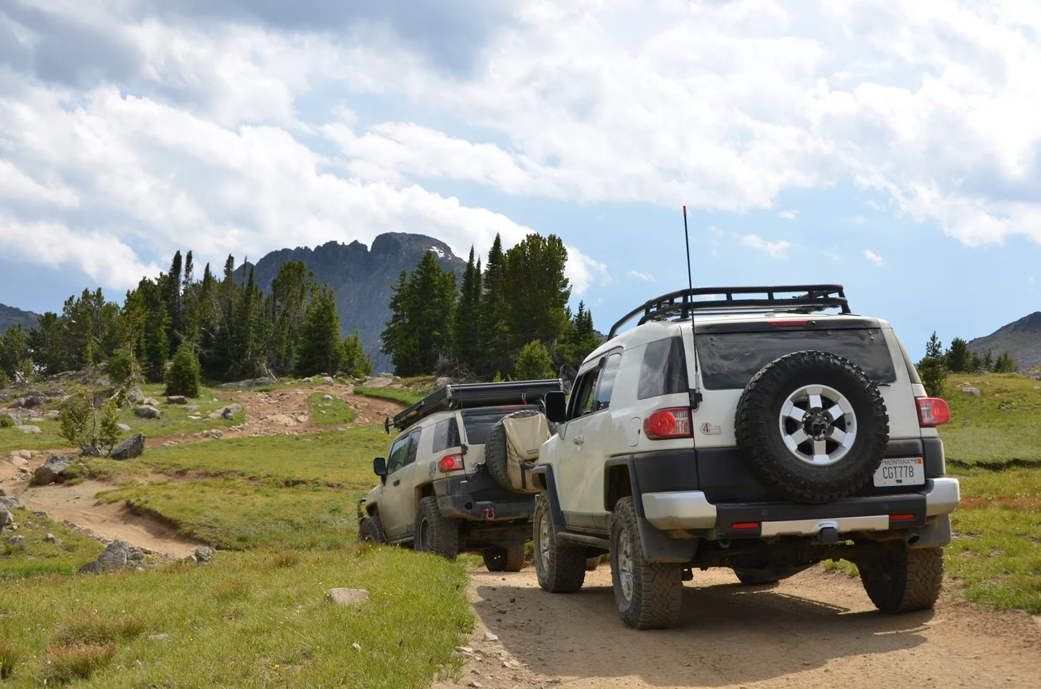 White Toyota FJ Cruiser with a spare tire and a tan FJ Cruiser parked on a meadow road with peaks behind on the Independence road above the Boulder River, Montana
