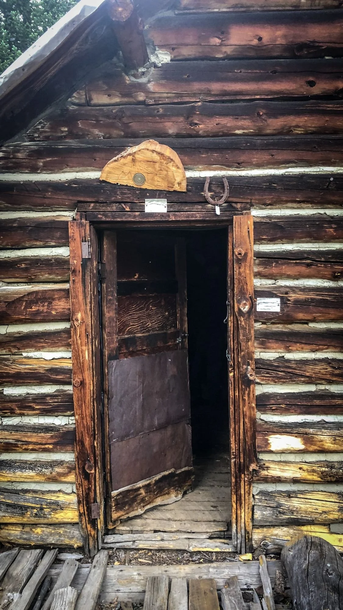 Weathered log cabin known as the No Tell Motel, with an open wooden door and a carved plaque above it on the North Meadow Creek trail to McKelvey Lake in the Tobacco Root Mountains, Montana