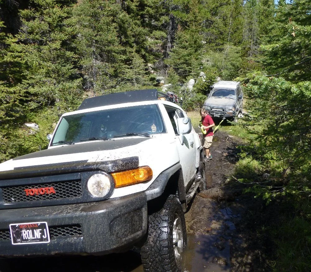 White FJ Cruiser in a muddy trail hole with another truck behind on a rocky forest trail in Montana