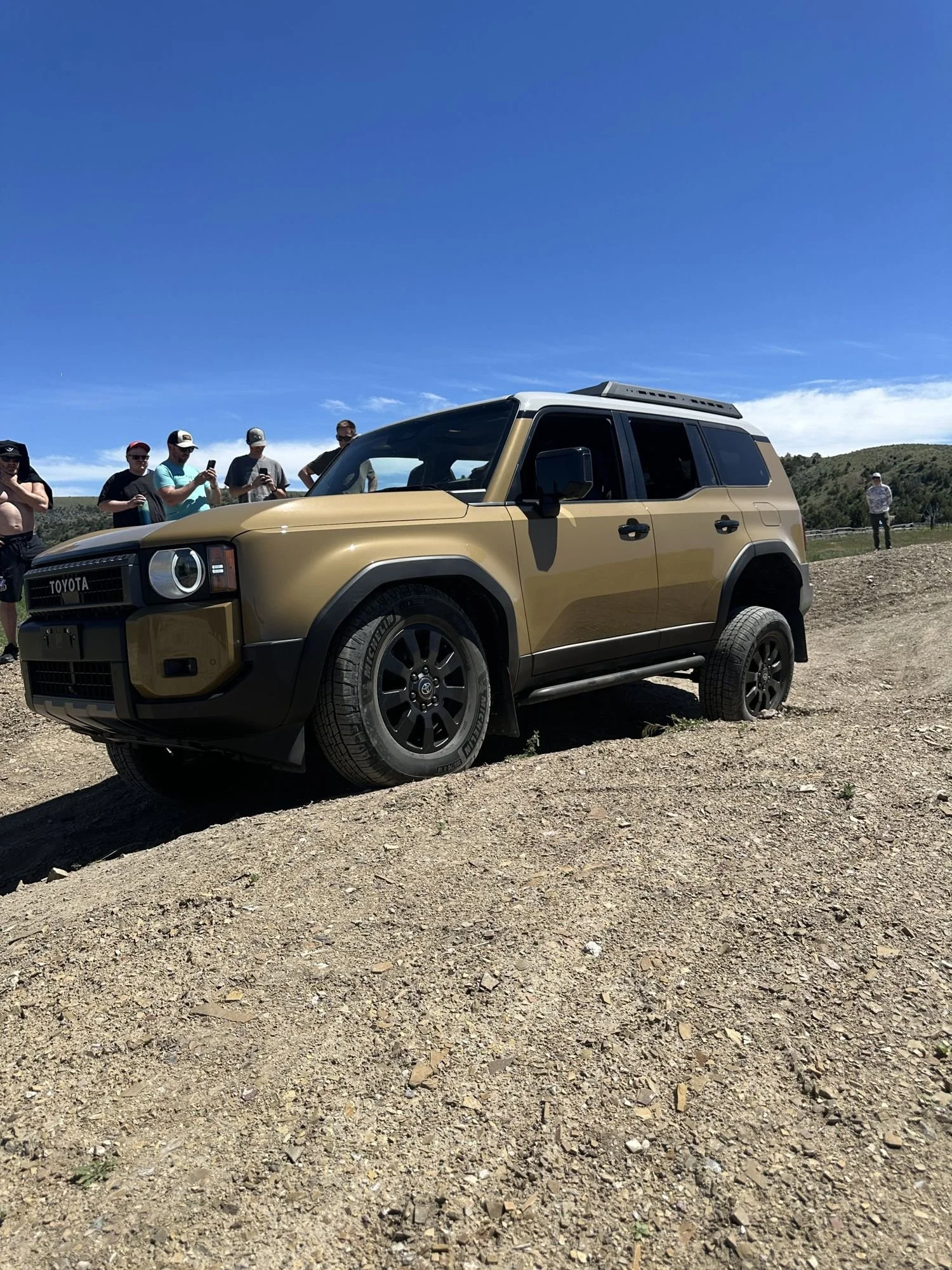 Tan Land Cruiser 250 tilted on an off-camber dirt slope while people watch from the top at XOverland Ranch near Bozeman, Montana