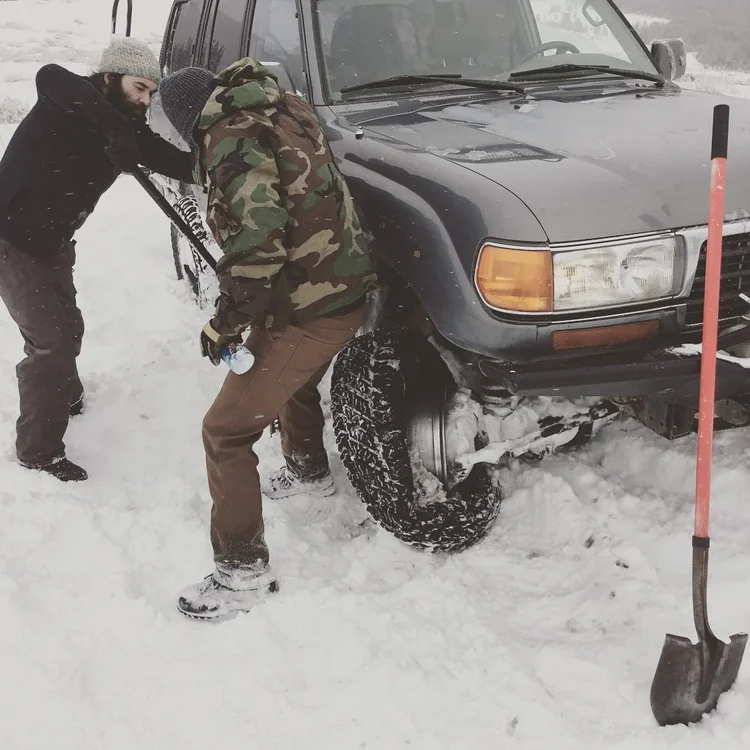 Two people crouched at the front tire of a gray Land Cruiser in falling snow, a shovel stuck upright beside them