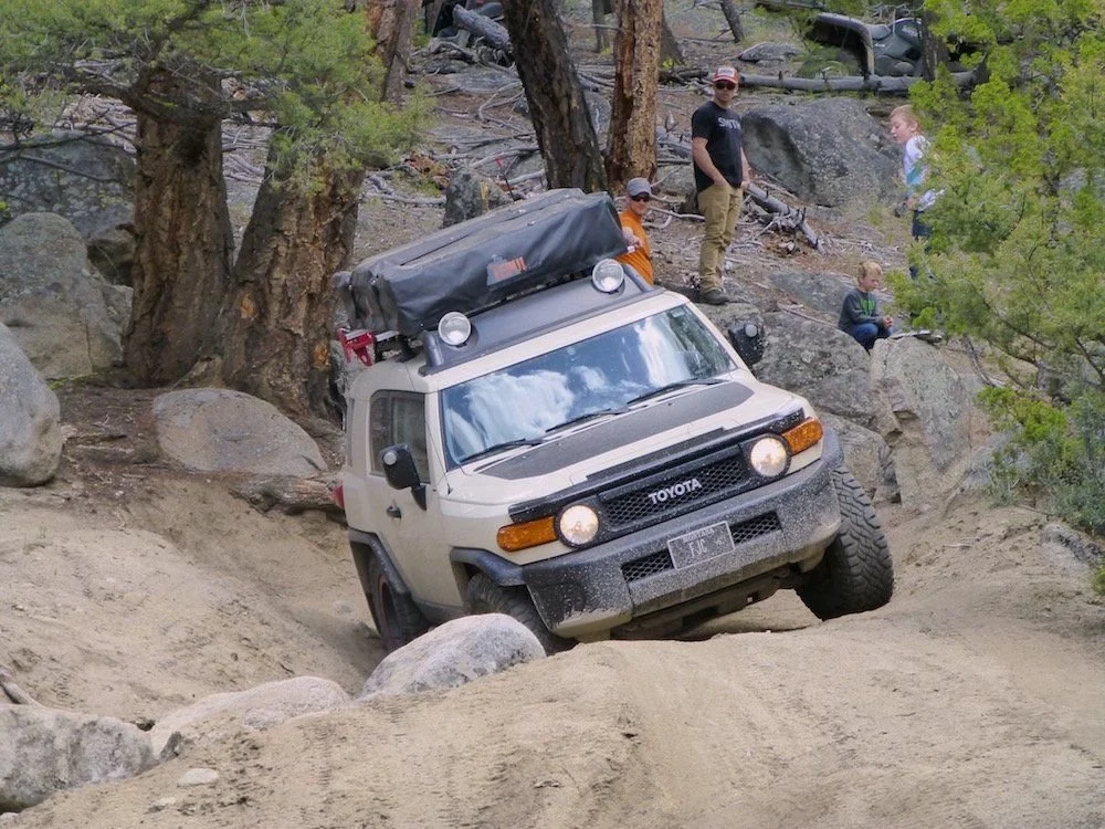 Tan Toyota FJ Cruiser crawling a sandy rock ledge while spotters watch from the boulders on the Ringing Rocks trail near Pipestone, Montana