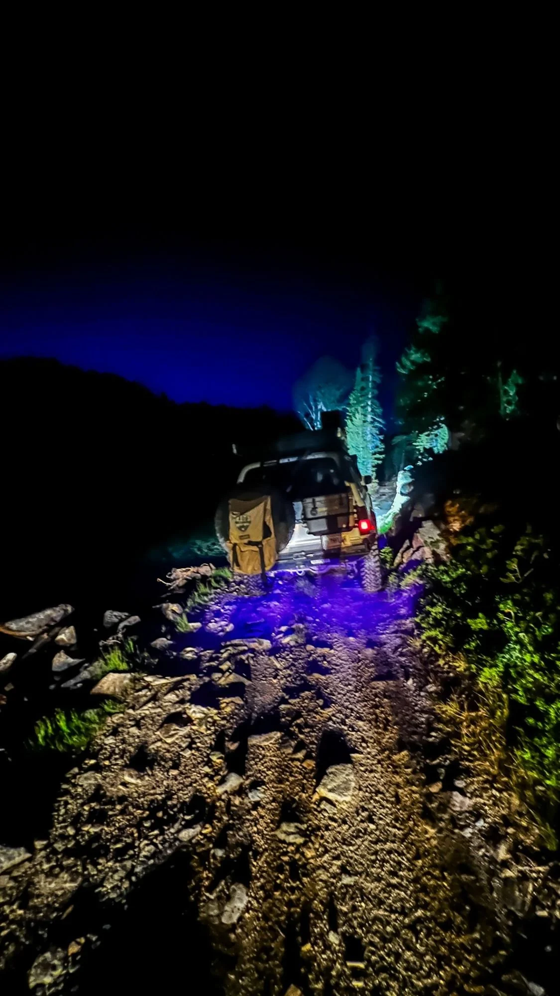 Truck on a rocky trail at night lit by blue rock lights under a dark blue sky near Cooke City, Montana