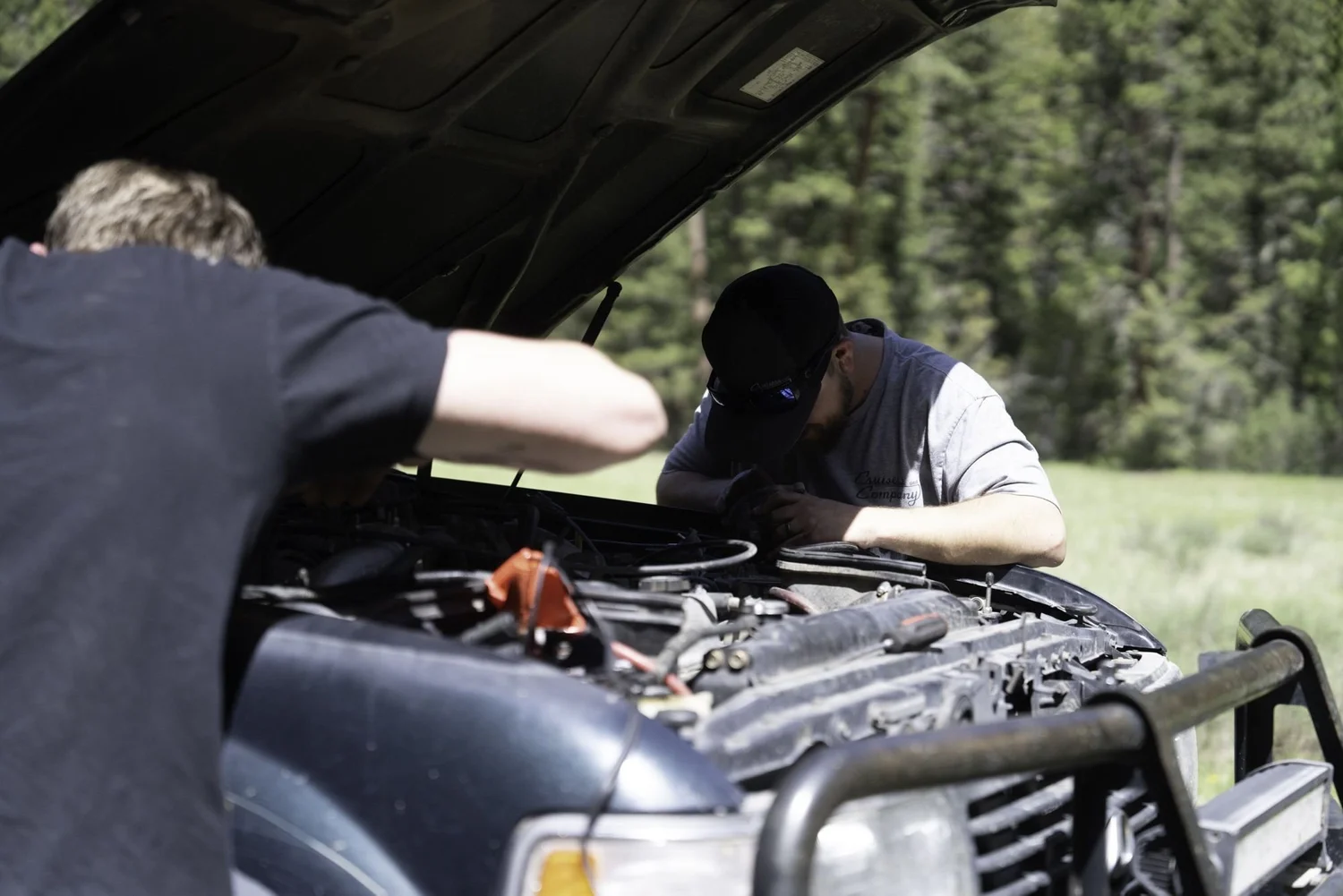 Two people leaning under the open hood of a blue truck working on the engine in a meadow on the Middle Fork Judith River road in the Little Belt Mountains, Montana
