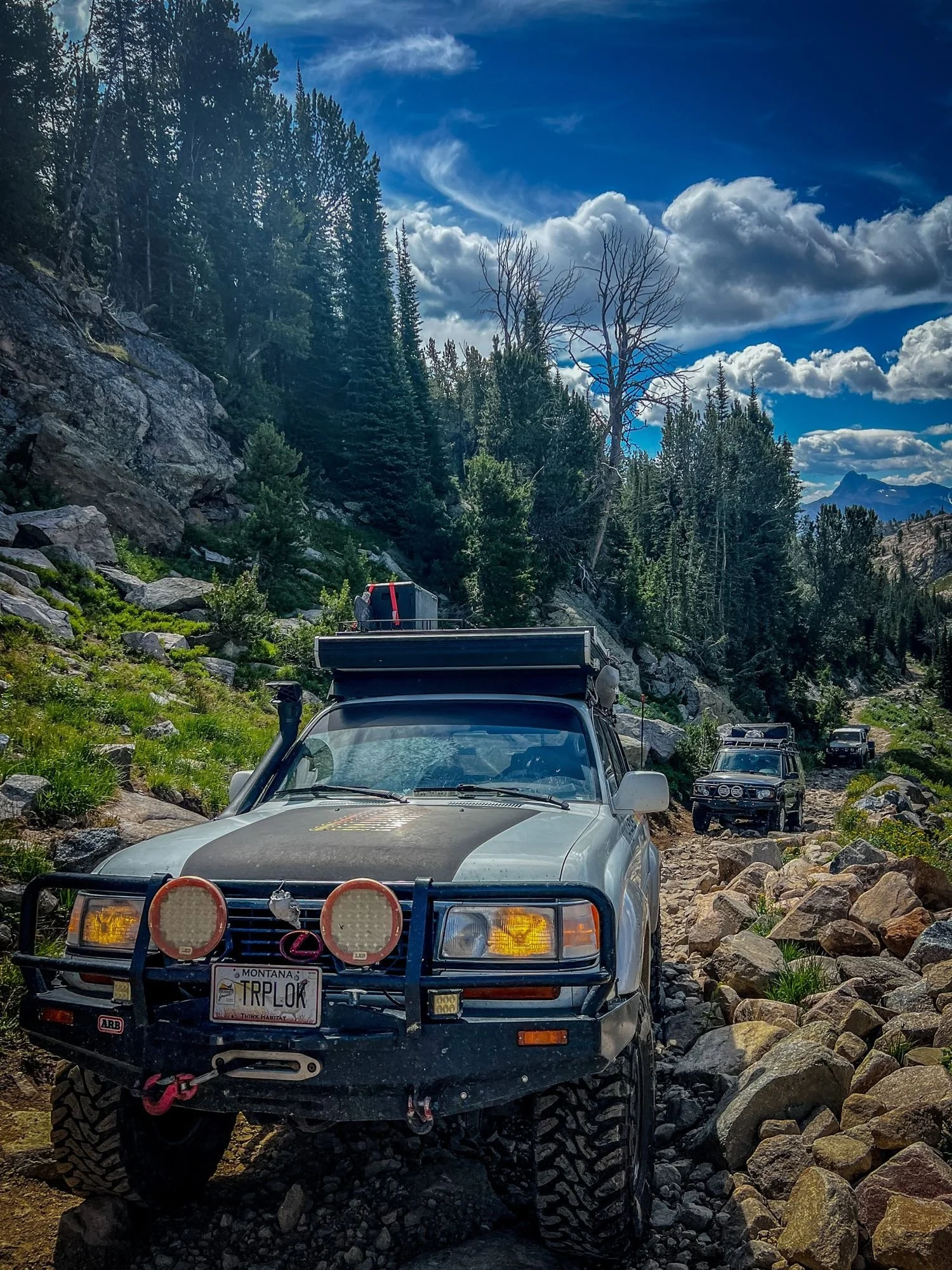 Silver Lexus LX450 with round auxiliary lights and a TRPLOK plate on a boulder-lined trail in the trees near Cooke City, Montana