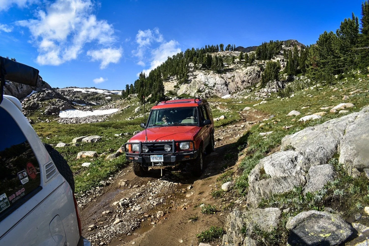 Red Land Rover Discovery climbing a rocky trail between granite boulders under a blue sky near Cooke City, Montana