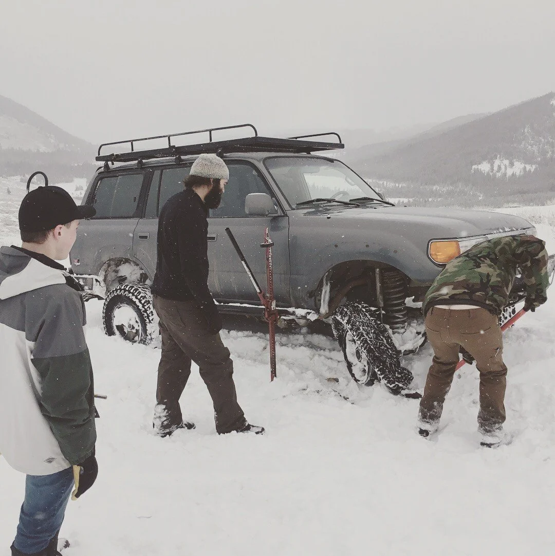 Three people working at a chained front tire of a gray Land Cruiser with a red Hi-Lift jack beside it during a December snow wheeling run in Montana