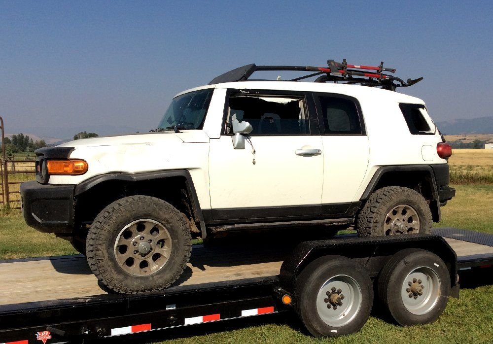 White FJ Cruiser with a broken side window loaded on a flatbed trailer in a sunny field on a rocky forest trail in Montana