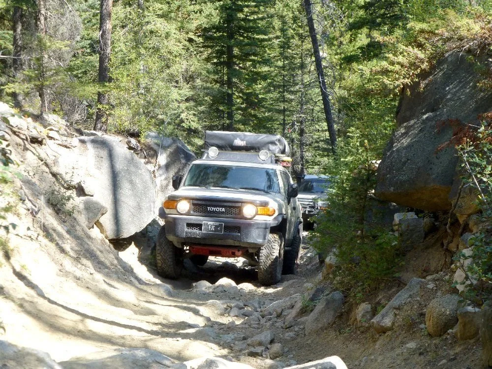 Tan FJ Cruiser with lights on climbing a narrow rocky chute between boulders on a rocky forest trail in Montana