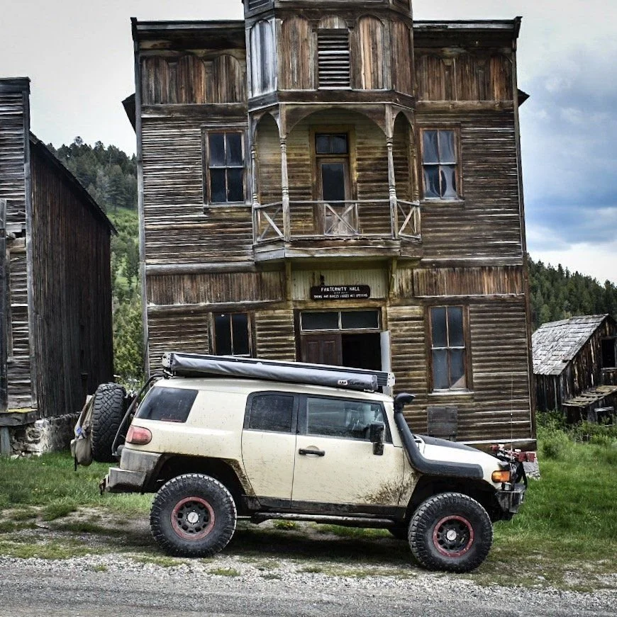 Tan FJ Cruiser with red wheels parked on gravel in front of a tall weathered wooden hall with a balcony in the Elkhorn ghost town near Boulder, Montana
