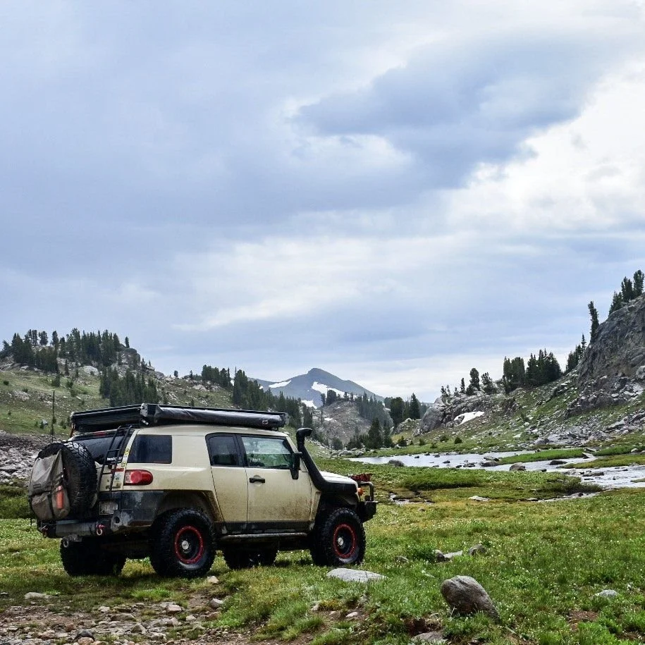Tan Toyota FJ Cruiser with a rear tire bag parked in a meadow beside a stream with peaks behind near Cooke City, Montana