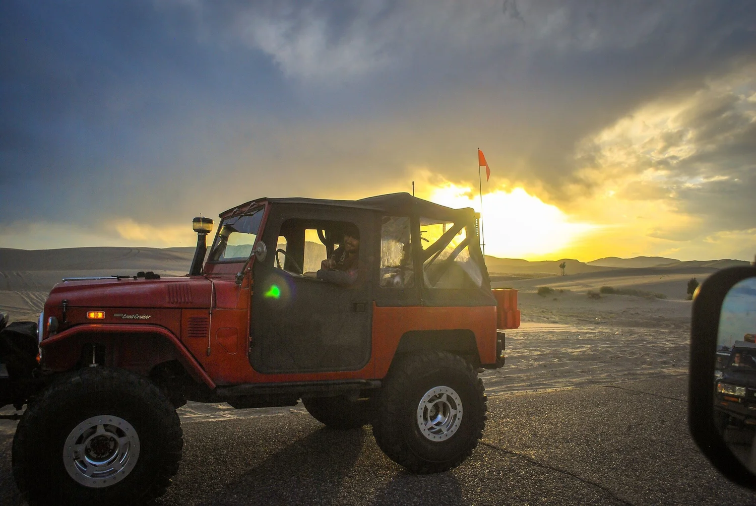 Red soft-top Toyota FJ40 Land Cruiser with a whip flag backlit by a stormy sunset at the St. Anthony Sand Dunes in Idaho