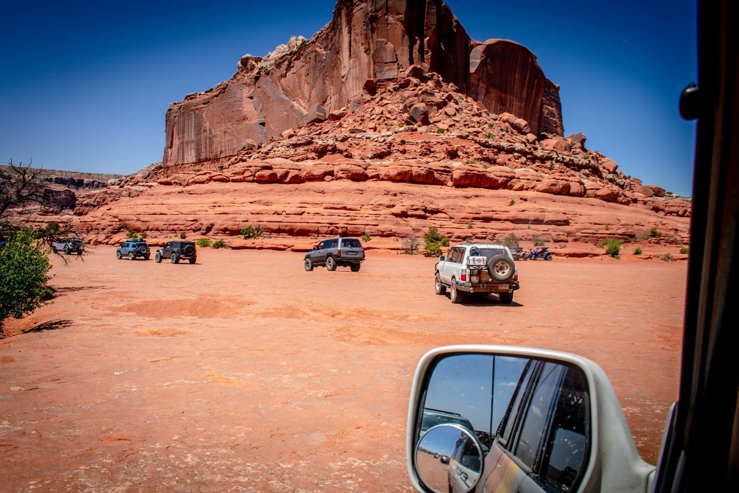 Group of trucks parked under a large red butte, seen from a truck window on the Seven Mile Rim trail near Moab, Utah