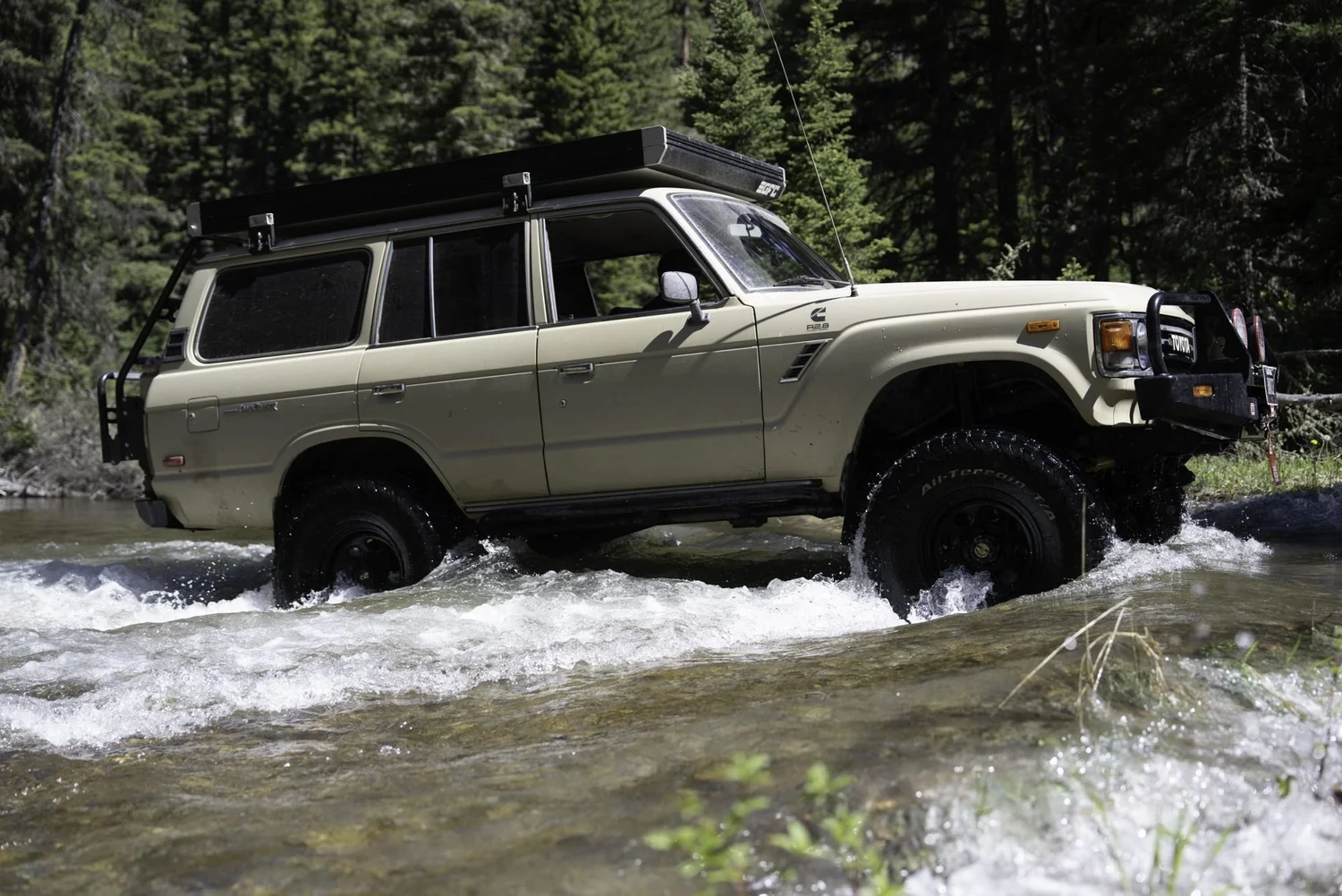 Tan Toyota Land Cruiser FJ60 with a roof rack pushing a wake through a river crossing on the Middle Fork Judith River road in the Little Belt Mountains, Montana