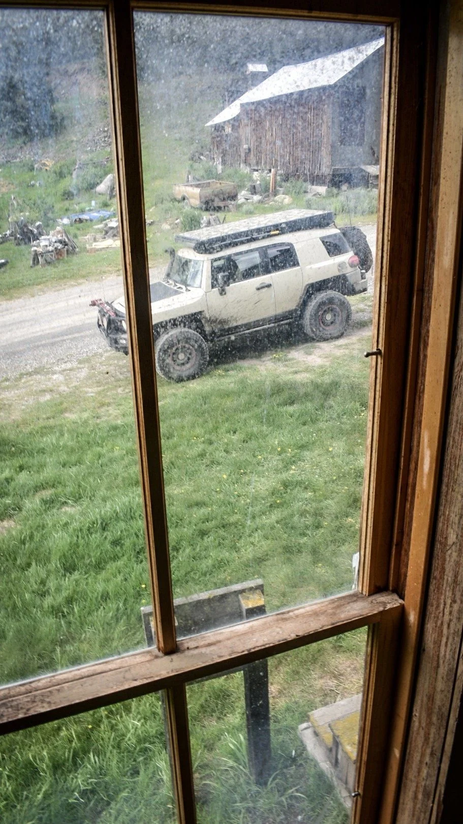 View through an old wooden window frame of a tan FJ Cruiser parked on the grass outside in the Elkhorn ghost town near Boulder, Montana