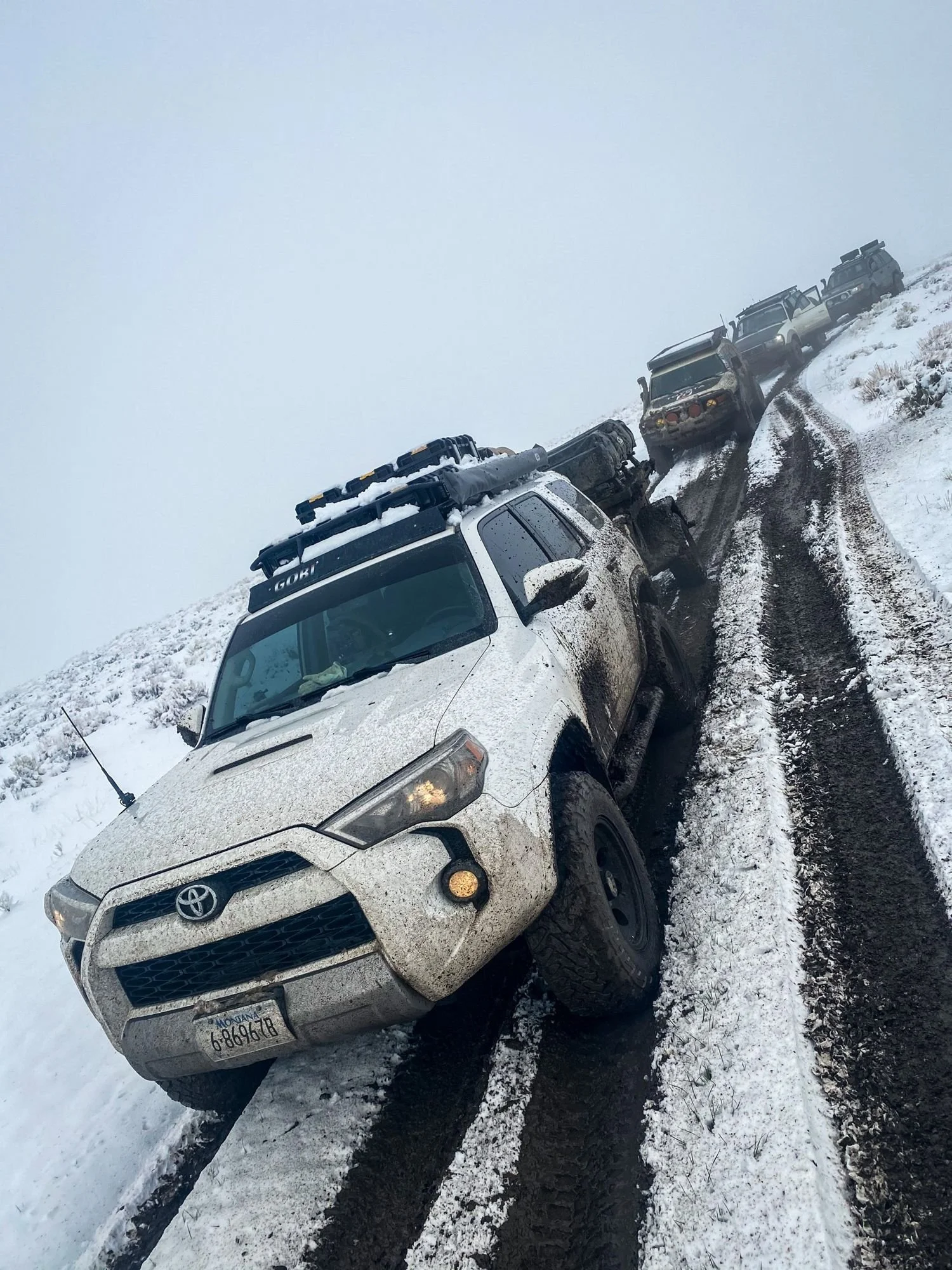 White Toyota 4Runner driving in deep, muddy ruts on a snowy hillside road with more trucks behind on a 406 Cruisers run in the upper Ruby River country, Montana
