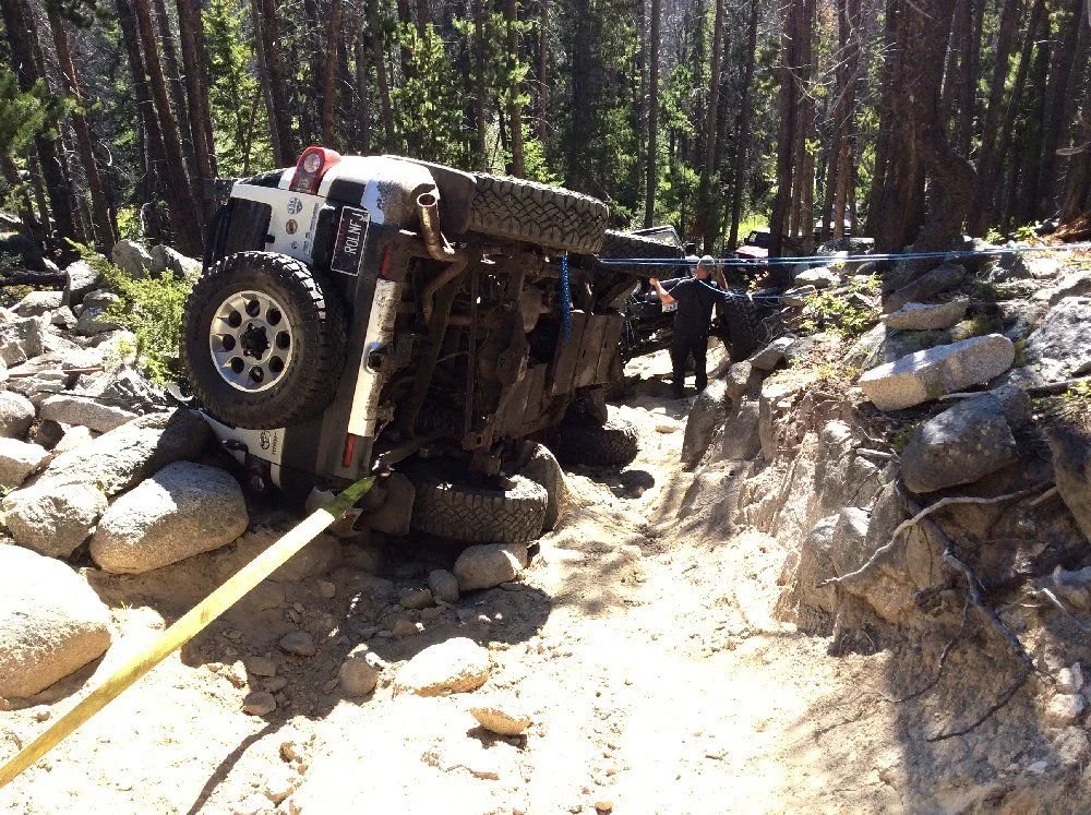 White Toyota FJ Cruiser lying on its side among boulders on a forest trail with a yellow recovery strap attached on a rocky forest trail in Montana