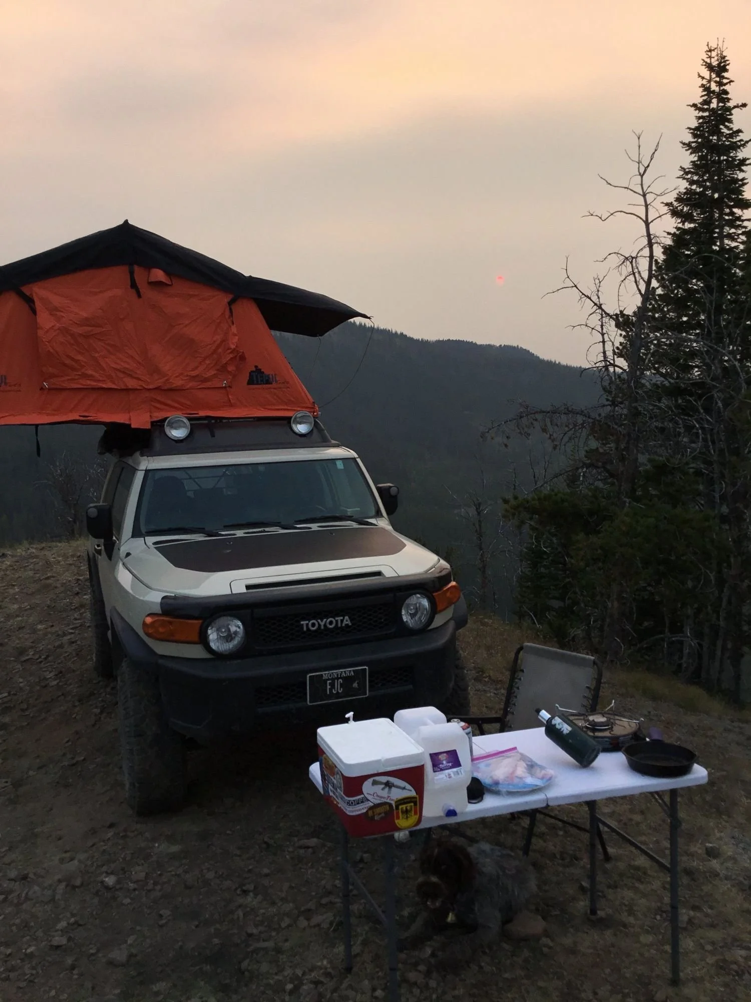 Tan FJ Cruiser with an orange rooftop tent on a ridgetop with a camp table, cooler and chair, and a dog lying under the table, in hazy light on Little Bear Road in the Gallatin Range near Bozeman, Montana
