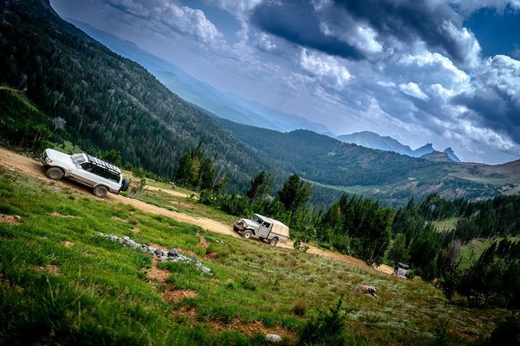 Land Cruisers on a narrow road cut into an alpine hillside above a valley near Cooke City.