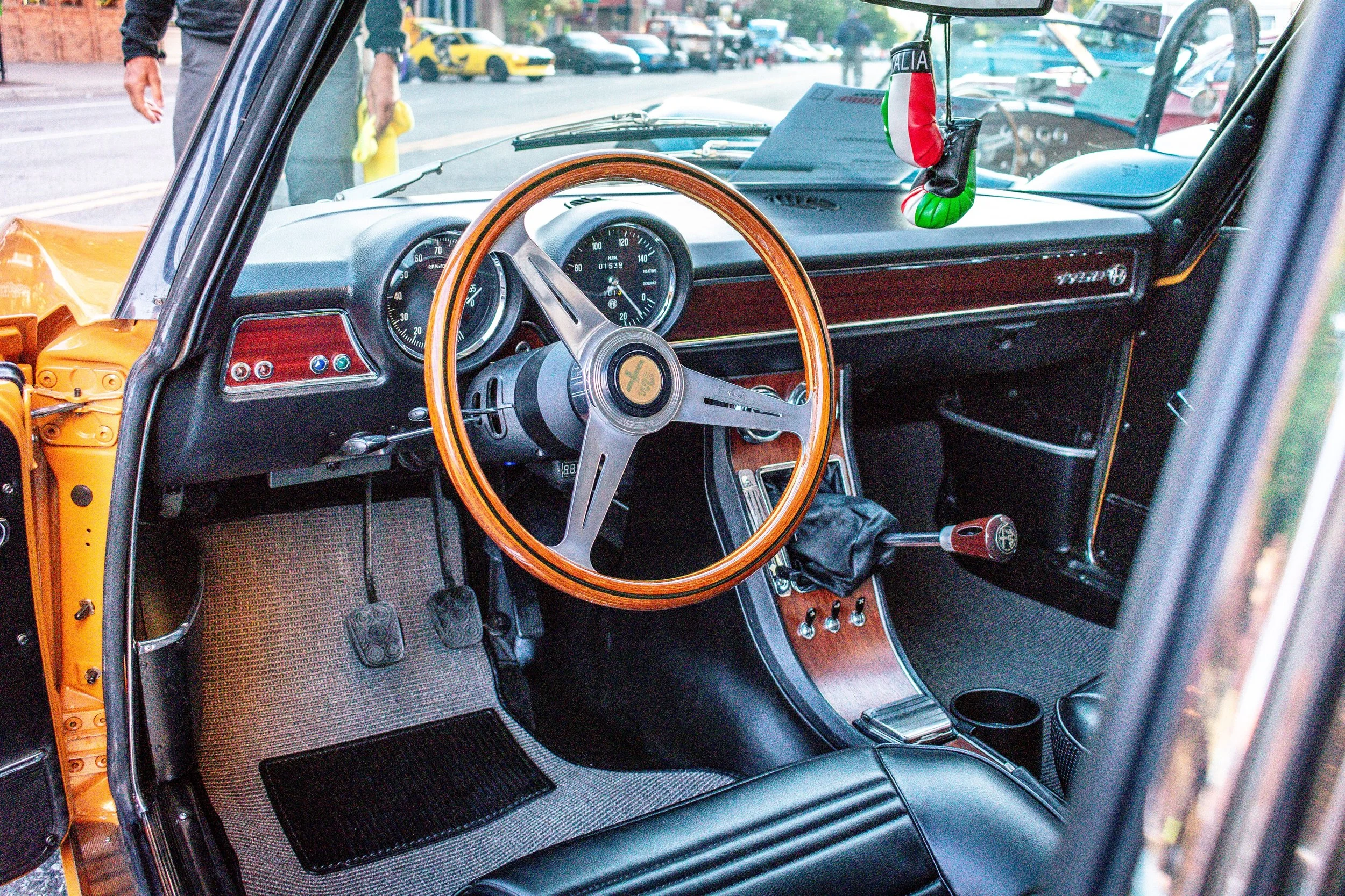 Interior of the Alfa Romeo GTV showing the wood-rimmed steering wheel, gated shifter, and an Italian flag ornament hanging from the mirror