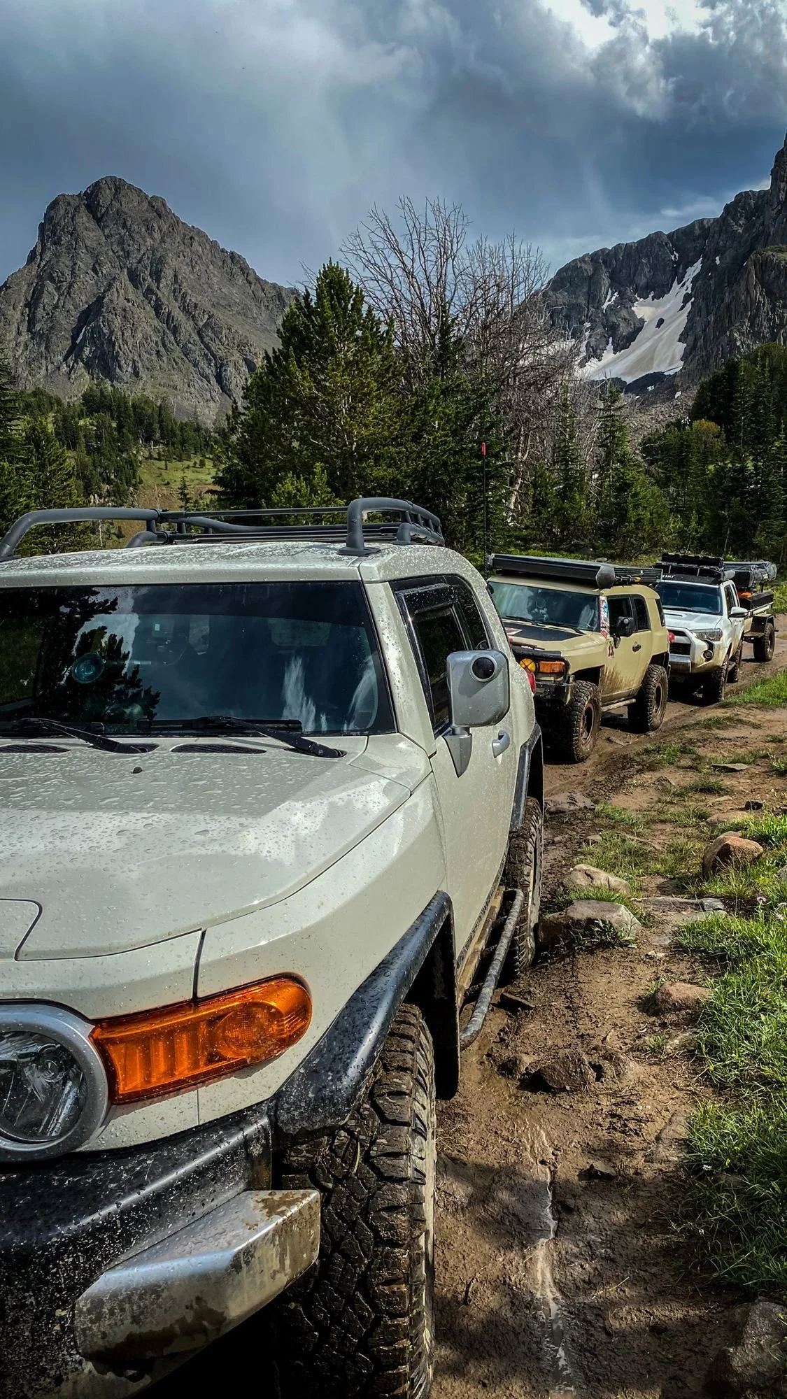 Line of FJ Cruisers on a muddy meadow road below a jagged peak on the Independence road above the Boulder River, Montana