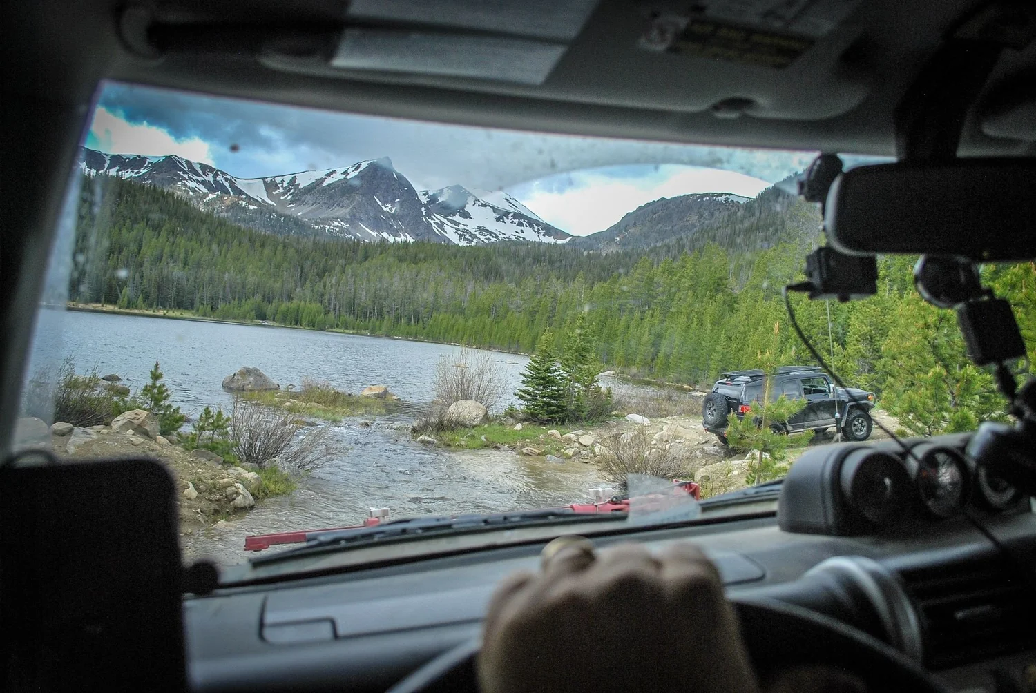 View through a windshield of a mountain lake with snowy peaks behind and a truck crossing a water channel on the North Meadow Creek Twin Lakes trail in the Tobacco Root Mountains, Montana