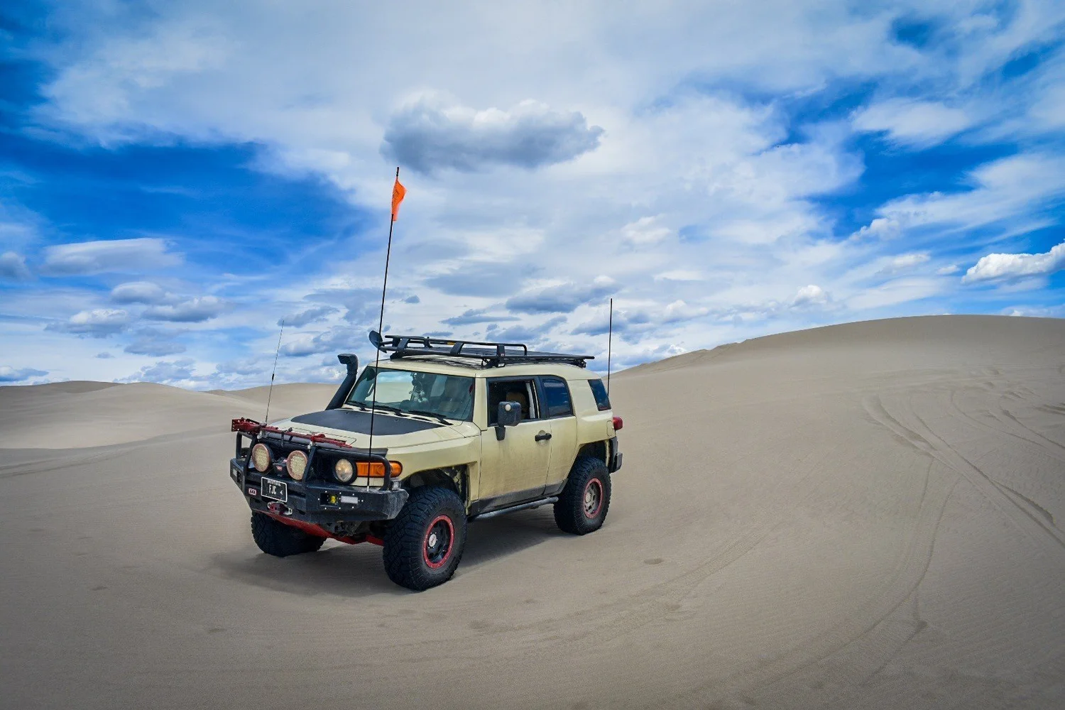 Tan Toyota FJ Cruiser with red wheels, a steel bumper, roof rack and whip flag on a dune under a blue sky at the St. Anthony Sand Dunes in Idaho