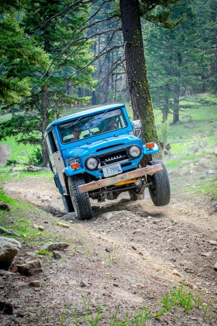 A blue FJ40 Land Cruiser with one front tire lifted off the ground on an uneven trail.
