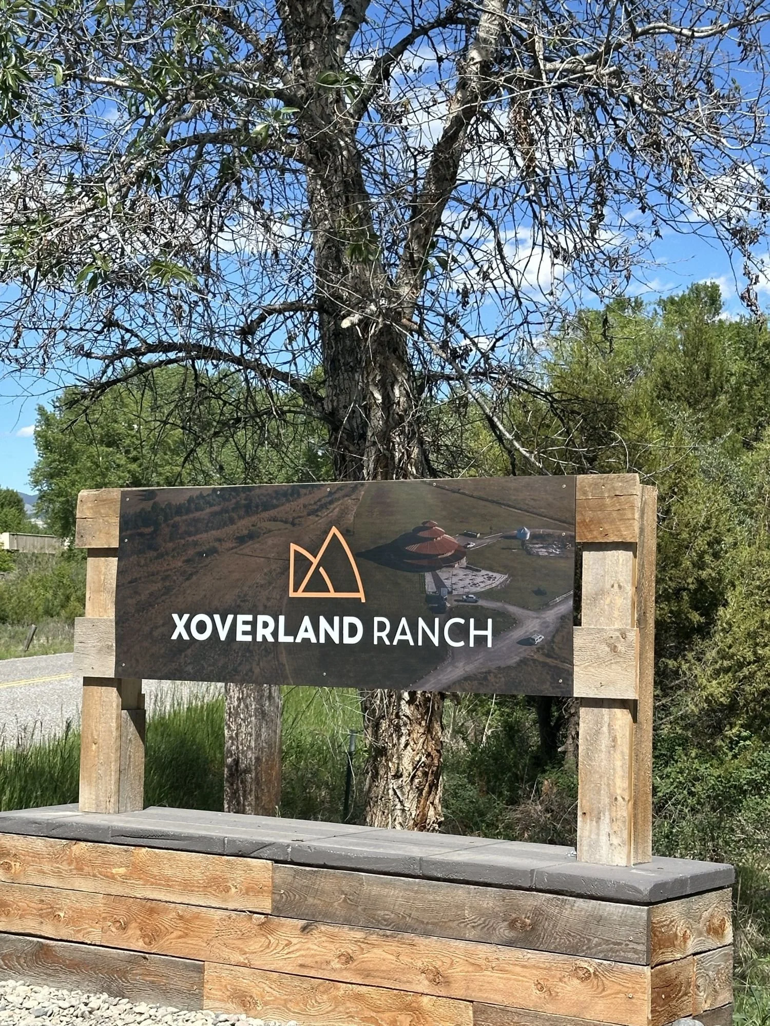 Wooden XOverland Ranch entrance sign with a mountain logo below a cottonwood tree near Bozeman, Montana