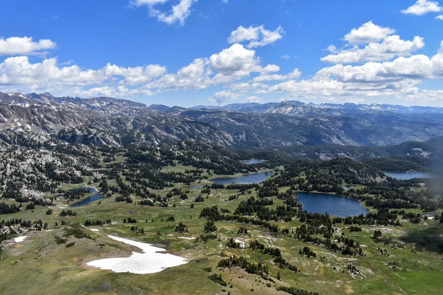 Wide view of a high plateau with several lakes, forest and distant mountains under puffy clouds near Cooke City, Montana