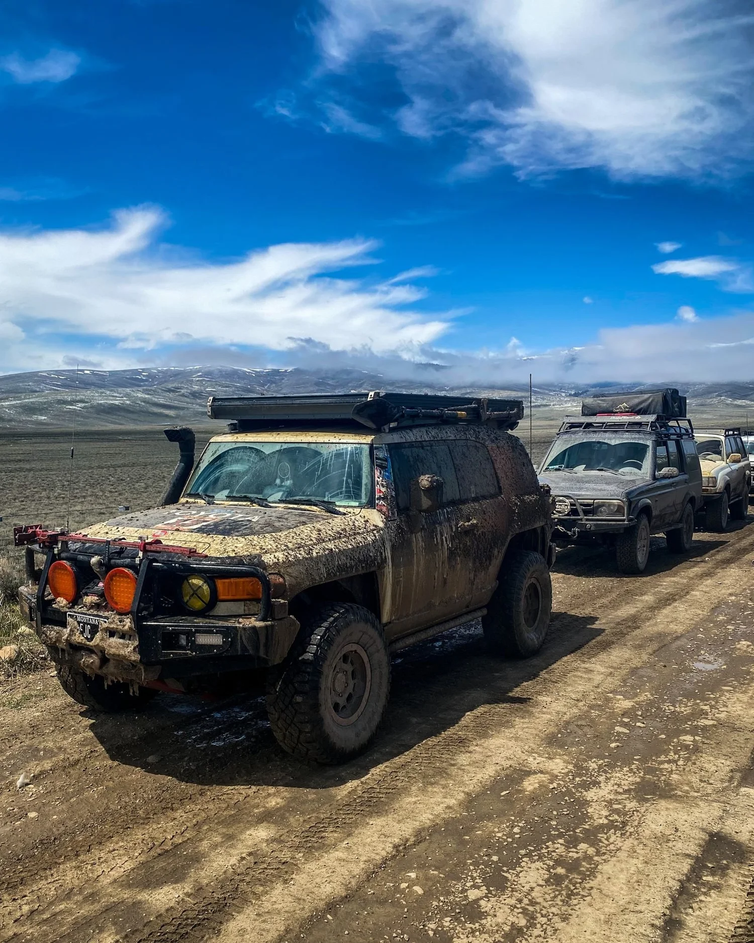 Mud-covered Toyota FJ Cruiser and another truck on a dirt road under a bright blue sky with wispy clouds on a 406 Cruisers run in the upper Ruby River country, Montana