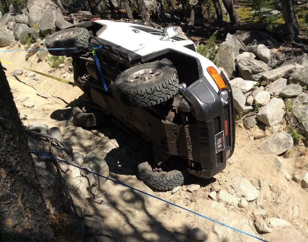 Underside of a white FJ Cruiser on its side with a winch line running uphill on a rocky forest trail in Montana