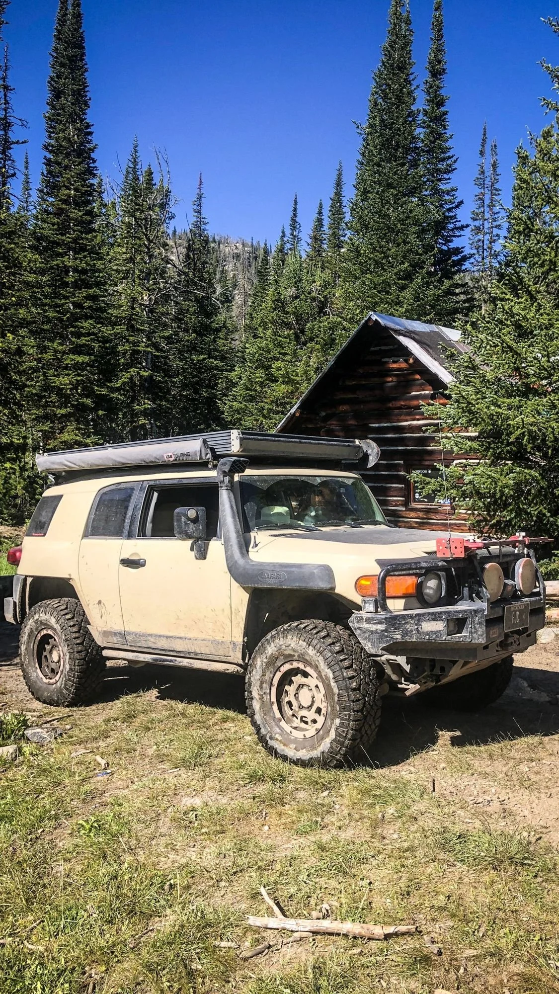 Tan FJ Cruiser parked in front of the No Tell Motel log cabin in a sunny clearing on the North Meadow Creek trail to McKelvey Lake in the Tobacco Root Mountains, Montana