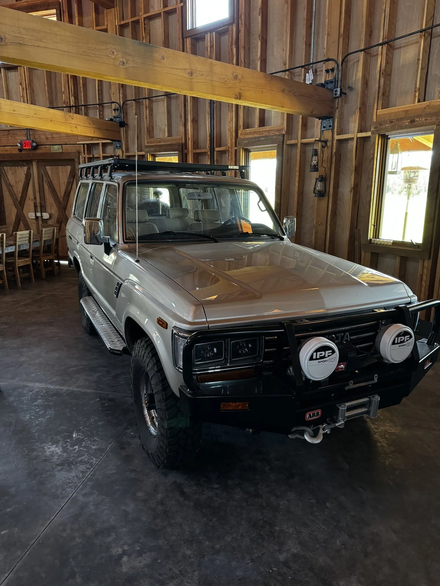 Silver Toyota Land Cruiser FJ62 with lights and a roof rack parked inside a wooden barn at XOverland Ranch near Bozeman, Montana