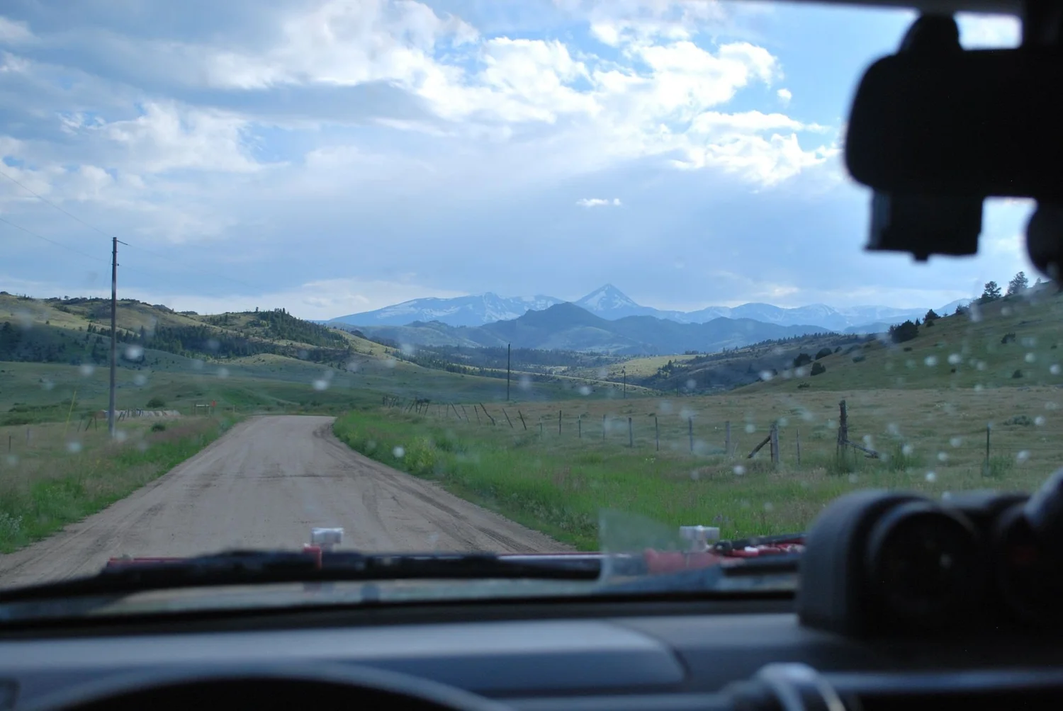 View through a windshield of a gravel road heading across open hills toward distant snowy mountains on the North Meadow Creek Twin Lakes trail in the Tobacco Root Mountains, Montana
