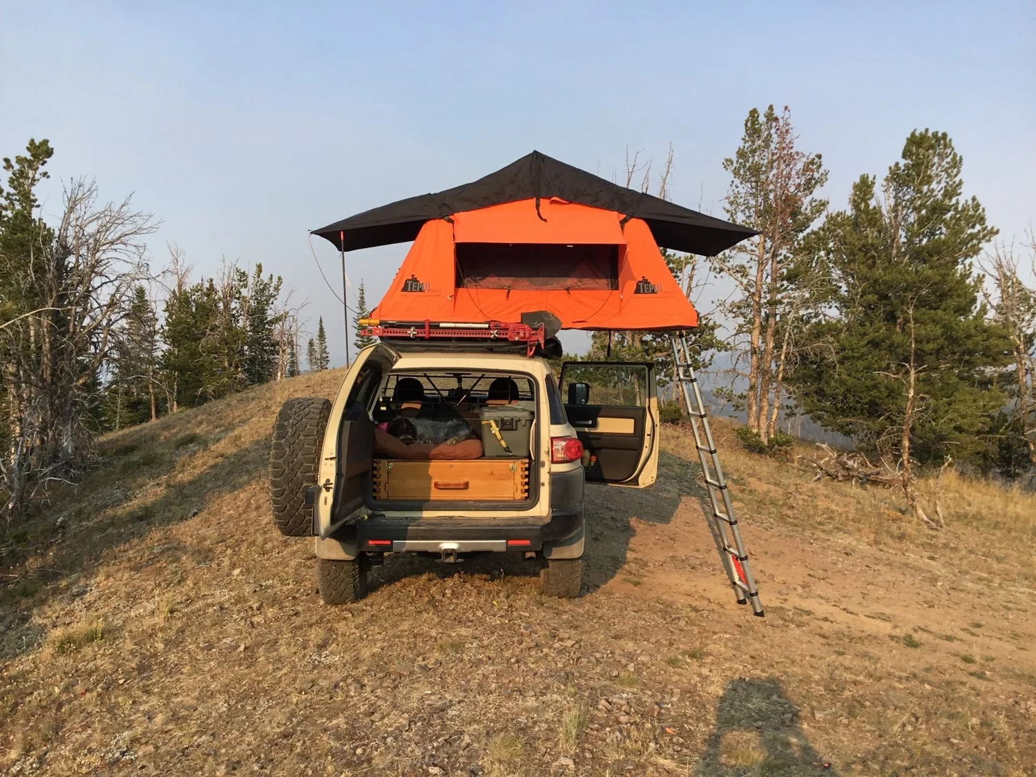 Tan FJ Cruiser with an orange rooftop tent and ladder parked on a grassy ridgetop under a hazy sky on Little Bear Road in the Gallatin Range near Bozeman, Montana