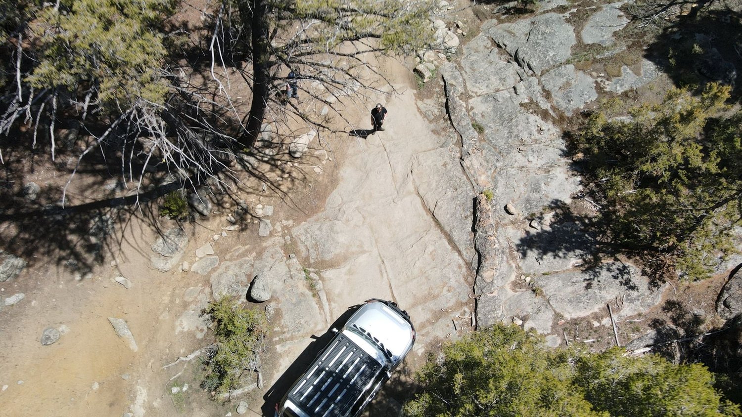 Overhead drone view of an off-road truck on a sloping granite slab with pine trees on the Ringing Rocks trail near Pipestone, Montana