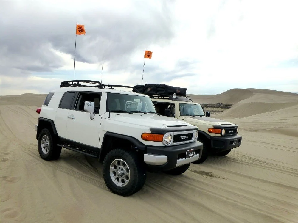 White and tan Toyota FJ Cruisers with orange whip flags parked on the sand at the St. Anthony Sand Dunes in Idaho