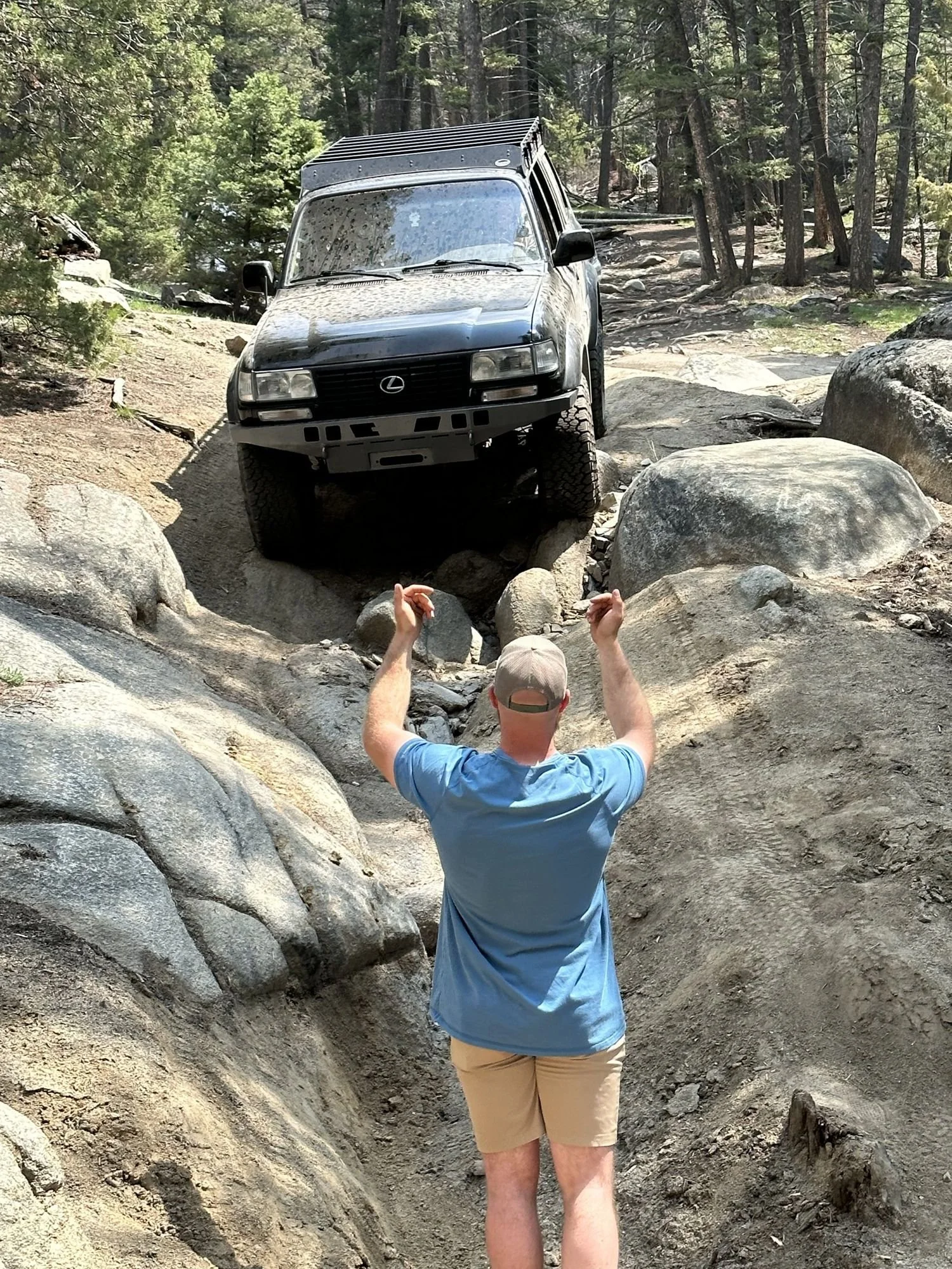Spotter in a blue shirt guiding a dark Toyota down a narrow granite boulder chute on the Ringing Rocks trail near Pipestone, Montana