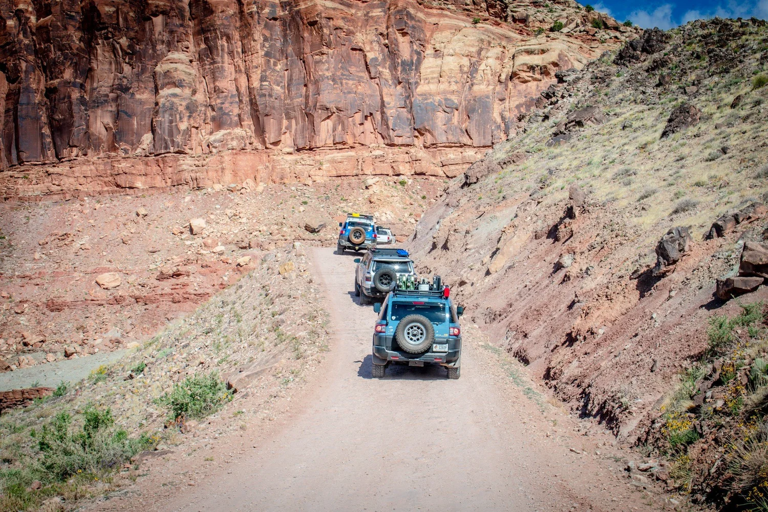 Three Land Cruisers single file on a dirt shelf road below a red cliff on the Seven Mile Rim trail near Moab, Utah