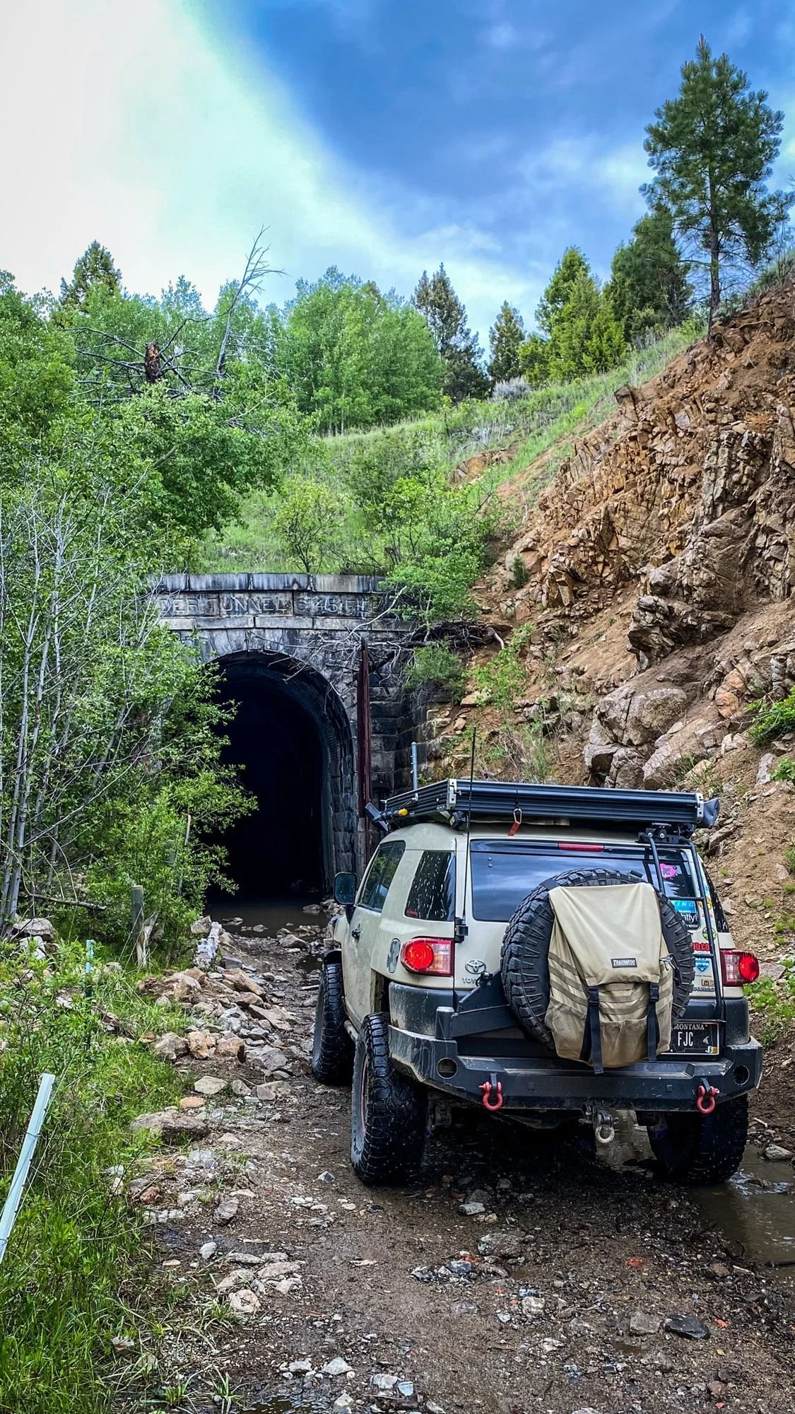 Tan Toyota FJ Cruiser with a rear tire bag parked on a rocky approach below the Wickes tunnel portal at the Wickes railroad tunnel near Jefferson City, Montana