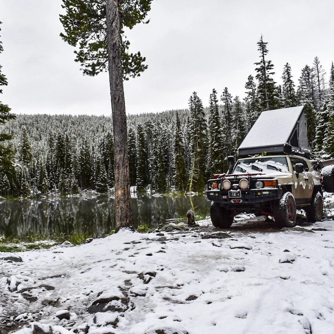 Tan FJ Cruiser with a rooftop tent parked beside a mountain lake after fresh snow in the Elkhorn Mountains near Boulder, Montana