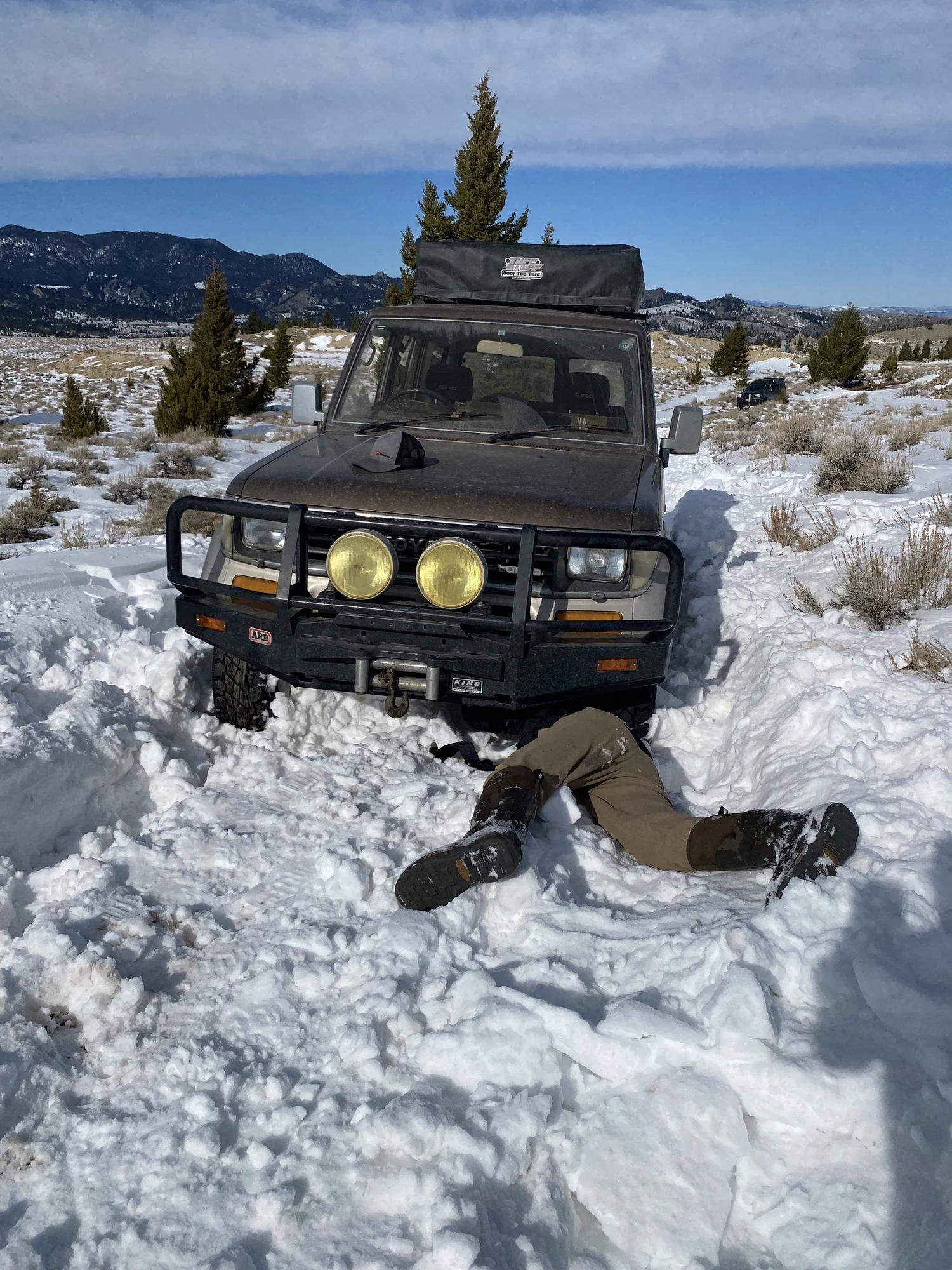 Person lying on their back in the snow digging out packed snow from under the front bumper of a Land Cruiser during a February snow wheeling run in Montana
