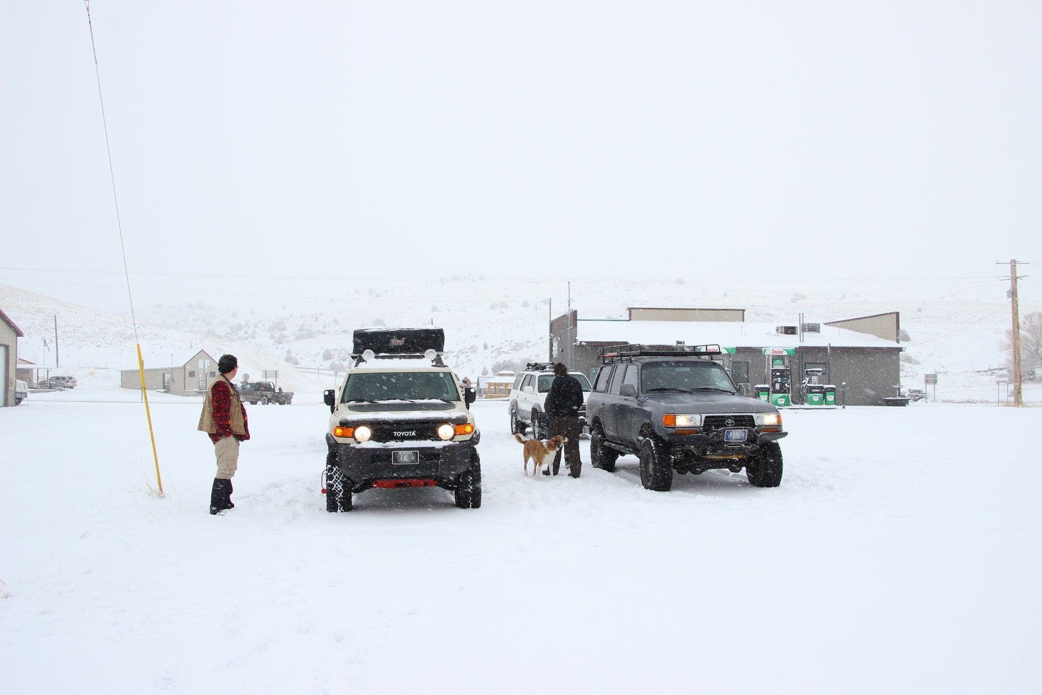 Toyota FJ Cruiser and an 80 Series Land Cruiser staged at a snowy small-town gas station in falling snow