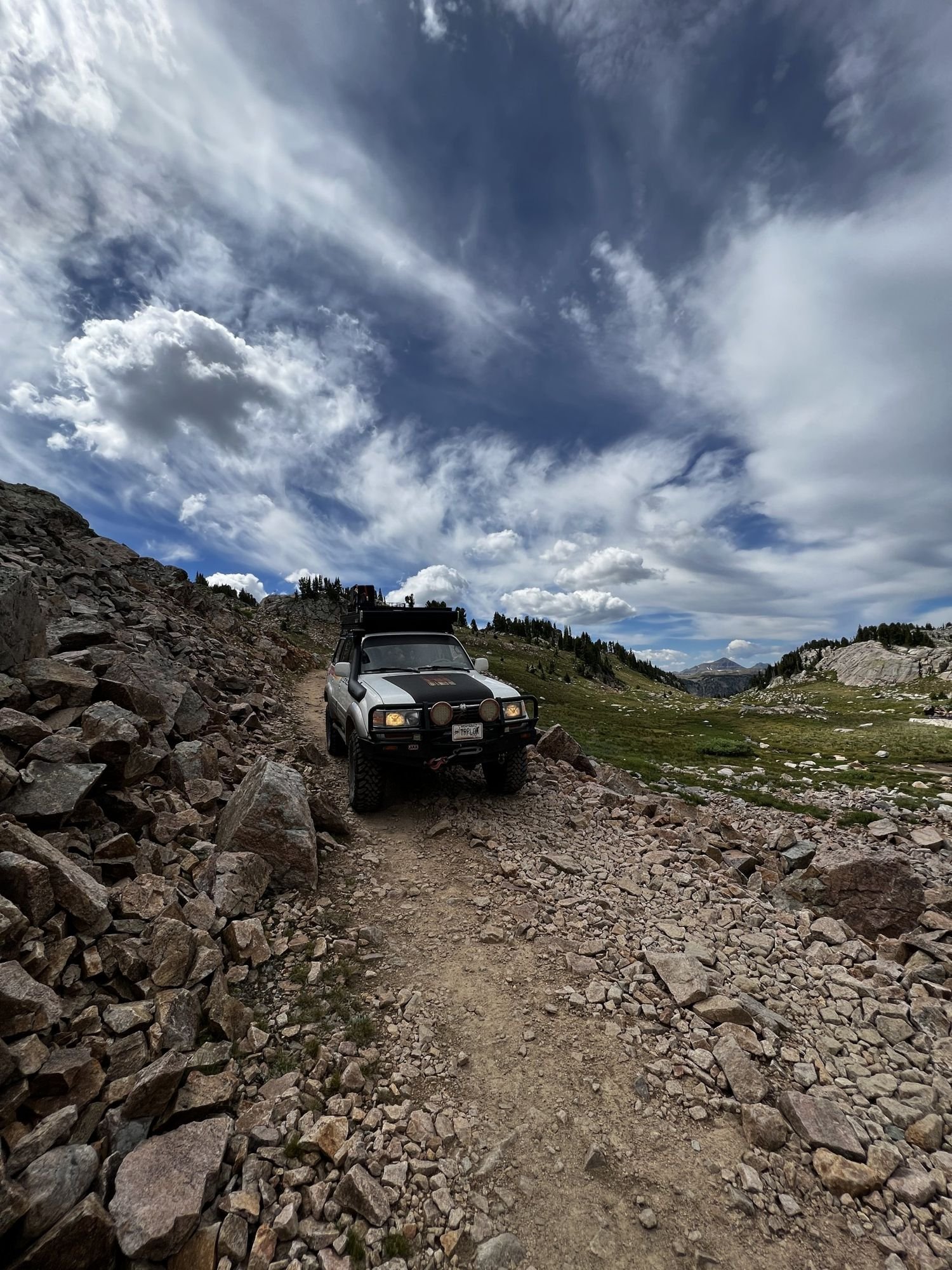 Rocky, boulder-strewn trail climbing a slope toward a truck under dramatic clouds near Cooke City, Montana