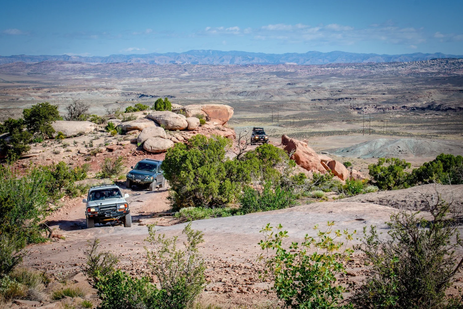 Three Land Cruisers on slickrock with mesas and distant mountains behind on the Seven Mile Rim trail near Moab, Utah