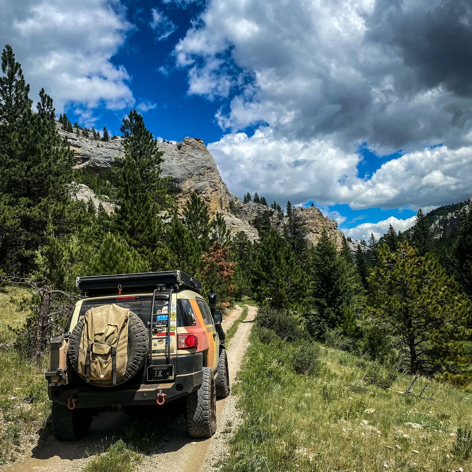 FJ Cruiser with a rear tire bag on a dirt two-track below limestone cliffs and big clouds on the Middle Fork Judith River road in the Little Belt Mountains, Montana