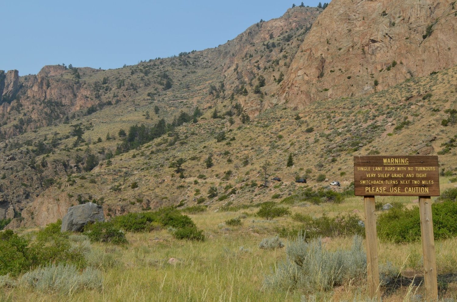 Brown warning sign at the base of a steep canyon slope warning of a single-lane road with a very steep grade and switchback turns on the Morrison Jeep Trail in Clarks Fork Canyon near Cody, Wyoming