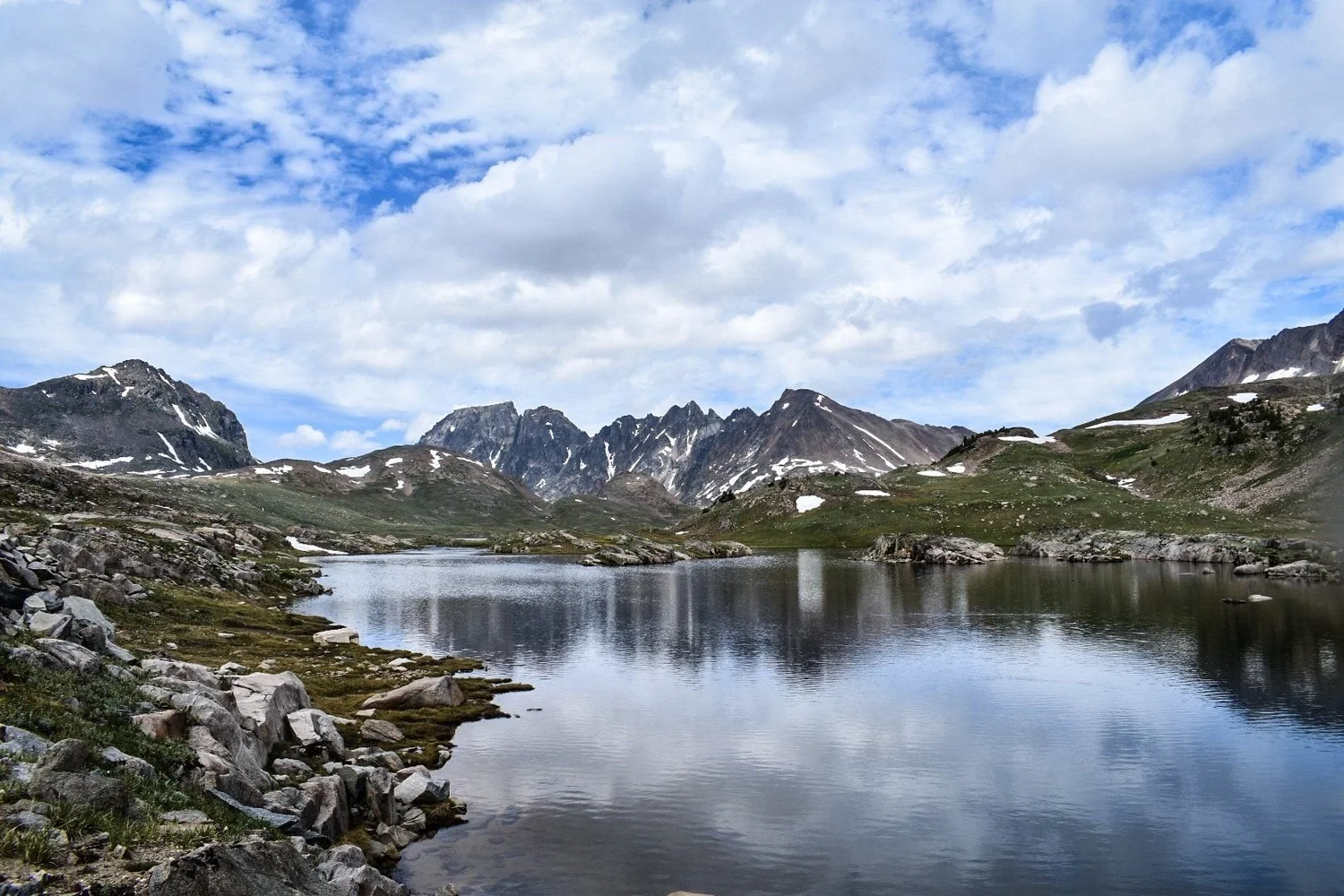 Alpine lake reflecting clouds and jagged peaks, with granite rocks in the foreground near Cooke City, Montana