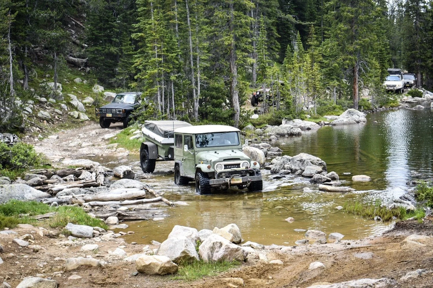 Green Toyota FJ40 Land Cruiser towing a small trailer through a rocky water crossing beside a pond near Cooke City, Montana